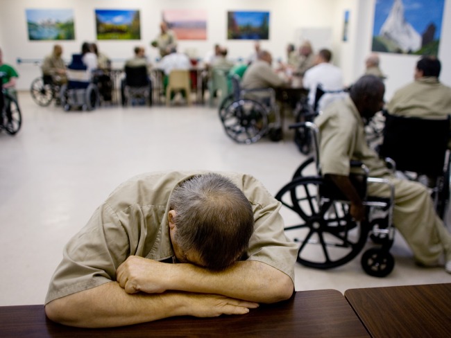 Elderly man in prison sits with his head on his desk. Behind him are several other elderly incarcerated men in wheel chairs.