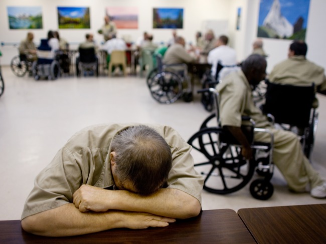 Elderly man in prison sits with his head on his desk. Behind him are several other elderly incarcerated men in wheel chairs.