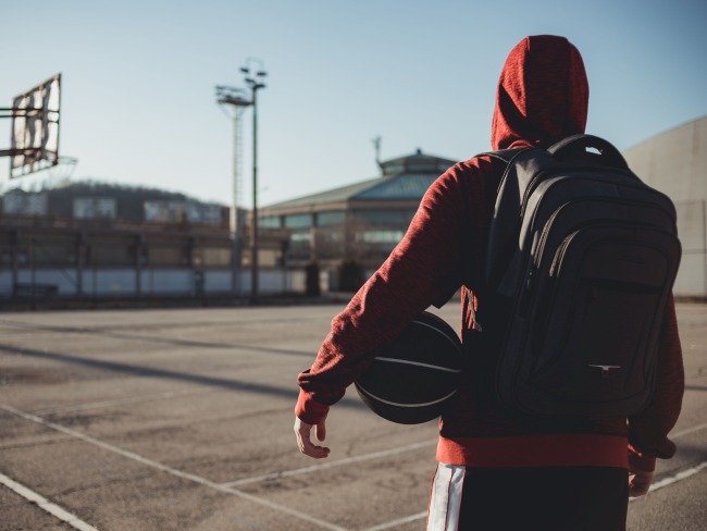Youth wearing red hoodie, black backpack, and holding a basketball on a basketball court