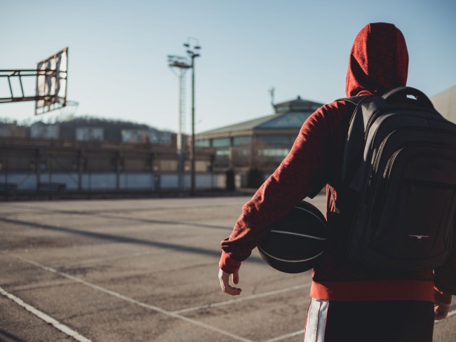 Youth wearing red hoodie, black backpack, and holding a basketball on a basketball court