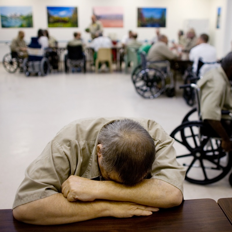 Elderly man in prison sits with his head on his desk. Behind him are several other elderly incarcerated men in wheel chairs.